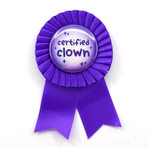 May include: A purple rosette ribbon with a circular center that reads "certified clown" in white and light blue. The center has a galaxy design with stars. The ribbon has two long, pointed tails.
