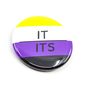 May include: A round button with the words "IT ITS" in black text on a white background. The button has a yellow, white, purple, and black striped design.