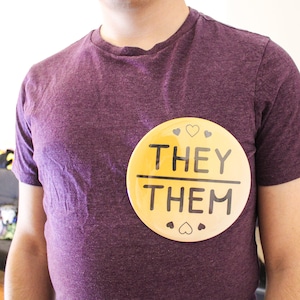 May include: A person wearing a purple t-shirt with a yellow button that says "They/Them" with small hearts around the edge.