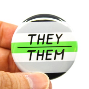 May include: A round button with a grey, white and green stripe pattern. The button has the text "THEY/THEM" in black.