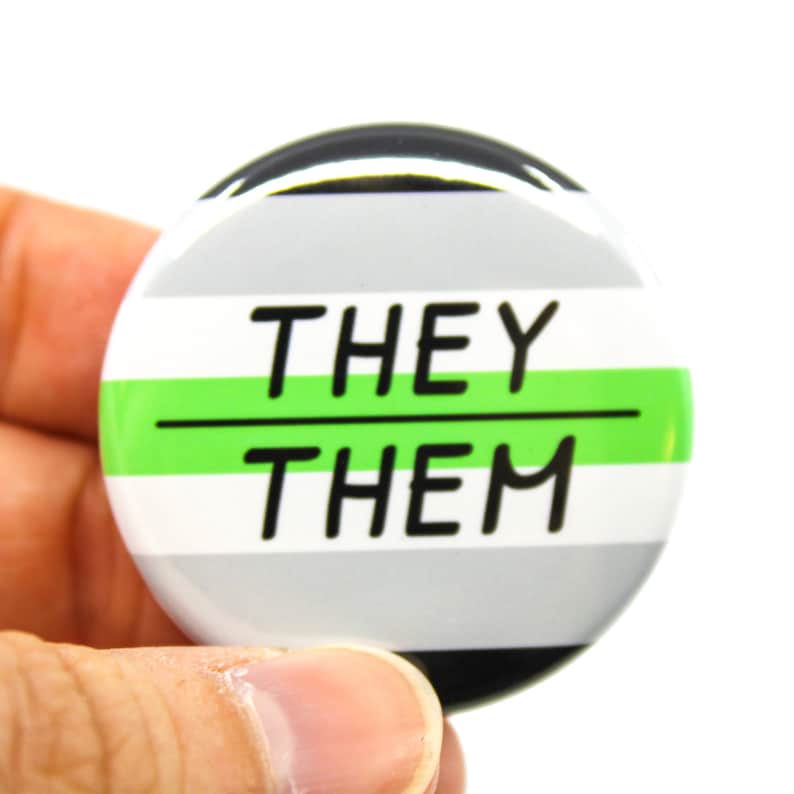 May include: A round button with a grey, white and green stripe pattern. The button has the text "THEY/THEM" in black.