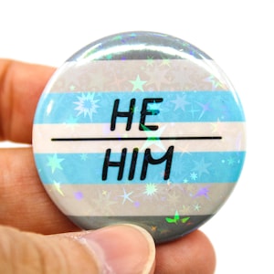 May include: A blue, white and grey button with a holographic finish. The button has a white stripe across the middle with the words "HE HIM" in black text.