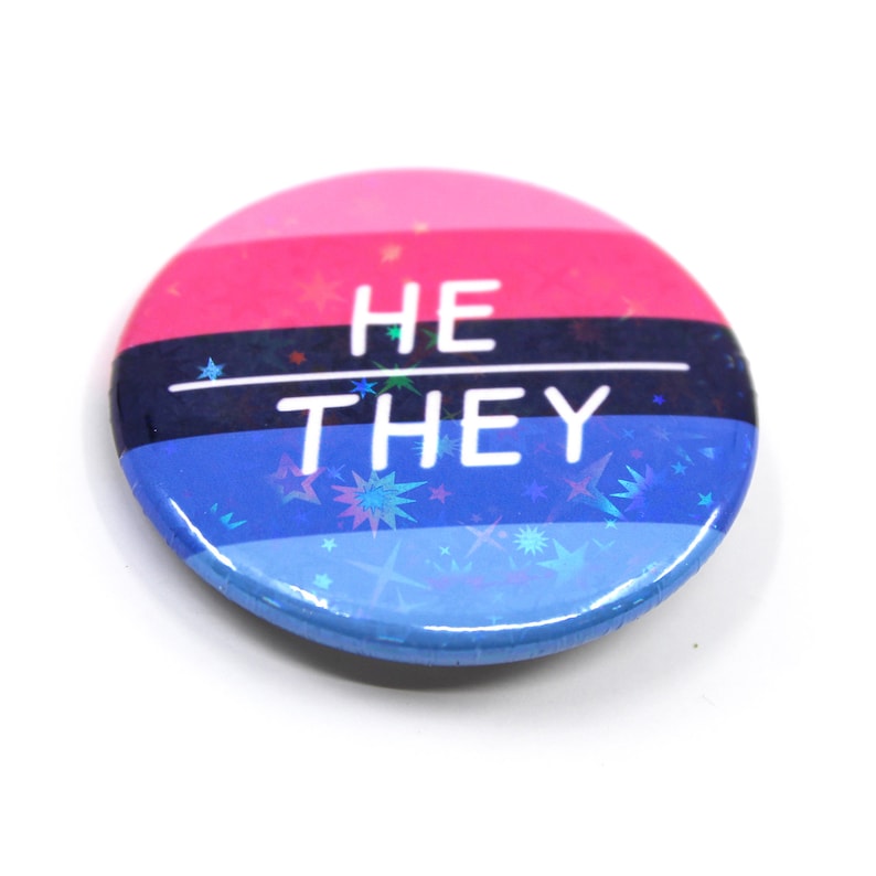 May include: A round button with a pink, blue and black striped background. The button has a holographic finish with stars and the text "HE THEY" in white.