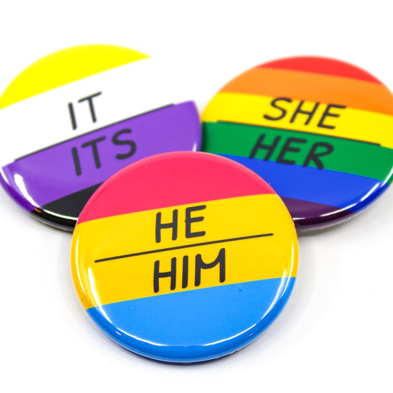 May include: Three round buttons with different pride flag colours and text. The first button is yellow, purple, black, and white with the text "IT ITS". The second button is red, yellow, blue, and light blue with the text "HE HIM". The third button is red, orange, yellow, green, blue, indigo, and violet with the text "SHE HER".