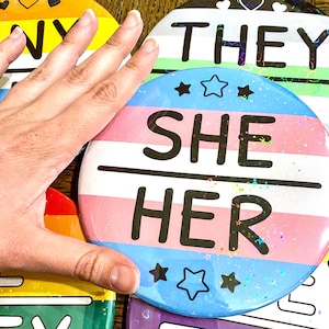 May include: A set of four round buttons with different pride flags and text. The buttons are blue, green, yellow, and pink with white stripes. The text on the buttons reads "They", "She/Her", "Gay", and "Bi".