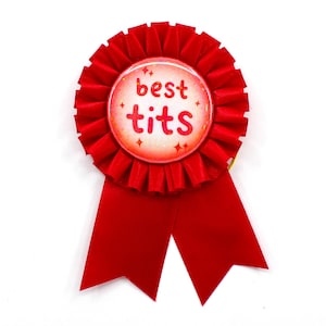 May include: A red rosette ribbon with a circular center. The center features the words "best tits" in red lettering, surrounded by small star accents. The ribbon has two long, pointed tails.