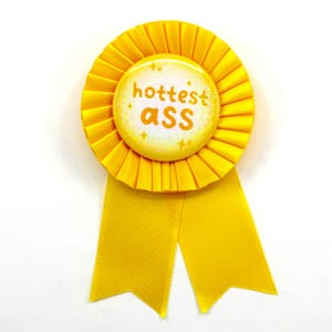 May include: A yellow rosette ribbon with a circular center badge. The badge has the words "hottest ass" in orange lettering. The rosette has a pleated design and two long ribbon tails.