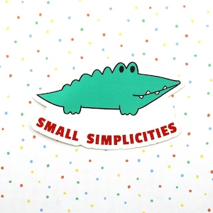 May include: A die-cut sticker featuring a cartoon green alligator with black eyes and white teeth. Below the alligator, the text "SMALL SIMPLICITIES" is printed in bold red capital letters against a white background.