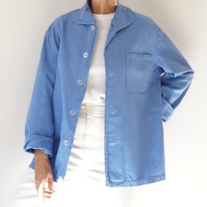 May include: A light blue jacket with a collared neckline and button-down front. The jacket has a single pocket on the chest and long sleeves. The model is wearing a white t-shirt and white pants.