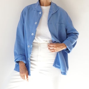 May include: A light blue button-down shirt with a chest pocket and rolled-up sleeves. The shirt is worn over a white t-shirt and paired with white trousers. The outfit is simple and casual, suitable for everyday wear.