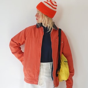 May include: An orange jacket with a dark collar, a navy blue shirt, white pants, and an orange and white striped beanie. A bright yellow bag is slung over the shoulder. The outfit consists of a zip-up jacket, a solid dark top, and light-colored trousers.