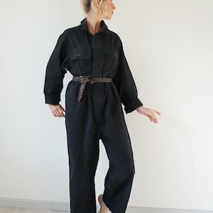 May include: A black jumpsuit with long sleeves, a collared neckline, and two chest pockets. The jumpsuit is cinched at the waist with a brown braided belt. The model is barefoot and standing in front of a white wall.