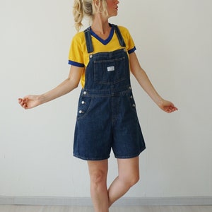 May include: Dark wash denim overall shorts with white button details. The shorts are paired with a yellow t-shirt with blue trim. The overall shorts have a front pocket and adjustable straps. The model is wearing the outfit.