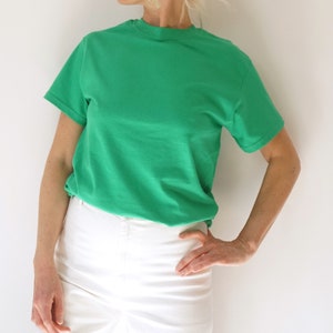 May include: A short-sleeved, solid green t-shirt paired with white pants. The t-shirt has a crew neck and a relaxed fit. The white pants are high-waisted. The outfit is simple and casual.