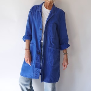May include: A long, cobalt blue work jacket with three large pockets and a button closure. The sleeves are rolled up, revealing a white t-shirt underneath. The jacket is worn over light blue jeans.