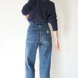 May include: Blue denim work pants with large back pockets and a Carhartt logo patch. The pants are paired with a navy blue sweatshirt. The style is casual and practical, suitable for everyday wear.