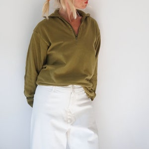 May include: Olive green long-sleeved top with a quarter-zip collar, paired with white trousers. The outfit is casual and appears to be made of a soft material. The clothing is simple and comfortable.