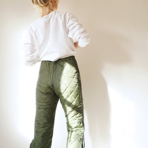 May include: A person wearing a white long-sleeved shirt and green quilted trousers. The trousers have a zip down the front and a diamond pattern.