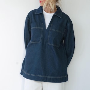 May include: A dark blue denim shirt with a V-neck collar and two square pockets on the chest. The shirt has long sleeves and a relaxed fit. The stitching is a contrasting light color. The model is wearing a white t-shirt underneath.
