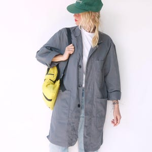 May include: A dark gray long-sleeve coat with a button-down front and two large patch pockets, worn over a white t-shirt and light blue denim jeans. A bright yellow backpack with black straps and a green baseball cap are also included.