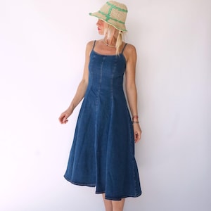 May include: A blue denim dress with spaghetti straps and a flared skirt. The dress has a fitted bodice and a midi length. The person is wearing a straw hat with green accents.