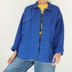 May include: A vibrant blue jacket with a classic collar, button-down front, and multiple pockets. The jacket is paired with light blue jeans and a yellow shirt underneath. The sleeves are cuffed, and the overall style is casual and functional.