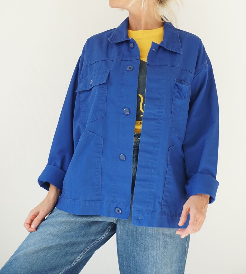May include: A vibrant blue jacket with a classic collar, button-down front, and multiple pockets. The jacket is paired with light blue jeans and a yellow shirt underneath. The sleeves are cuffed, and the overall style is casual and functional.