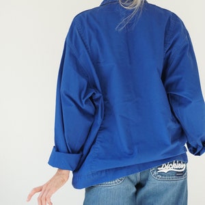 May include: A cobalt blue jacket with rolled-up sleeves. The jacket is paired with light-wash denim jeans. The back pocket of the jeans features the Dickies logo in white script.