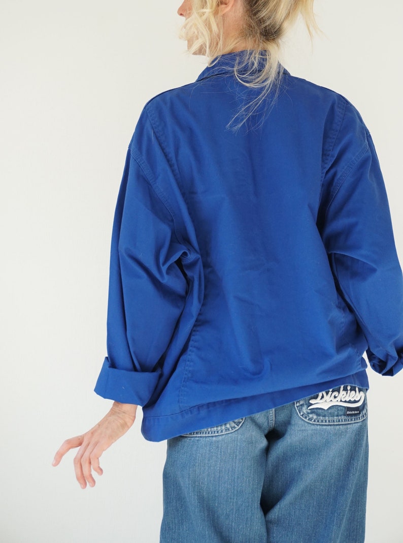 May include: A cobalt blue jacket with rolled-up sleeves. The jacket is paired with light-wash denim jeans. The back pocket of the jeans features the Dickies logo in white script.