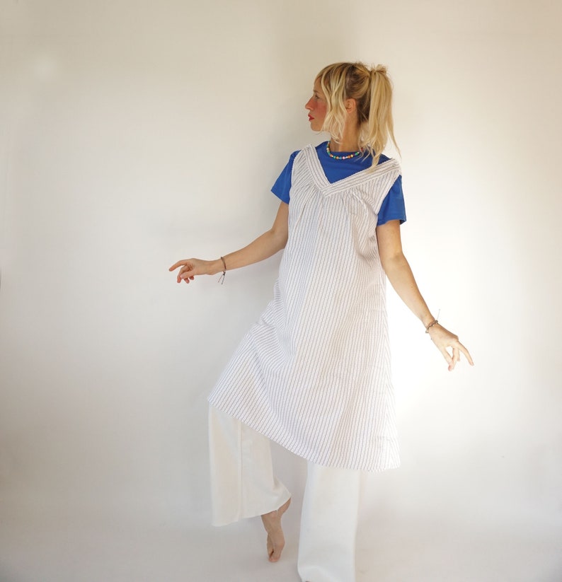 May include: A white and blue striped dress with a V-neckline and a loose fit. The dress is worn over a blue t-shirt and white trousers.