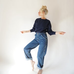 May include: A navy blue crew neck sweatshirt and blue denim jeans. The jeans have a classic fit with a straight leg and rolled cuffs. The model is wearing the outfit in front of a white background.
