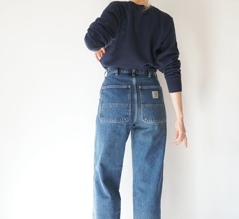 May include: Blue denim work pants with large back pockets and a Carhartt logo patch. The pants are paired with a navy blue sweatshirt. The style is casual and practical, suitable for everyday wear.