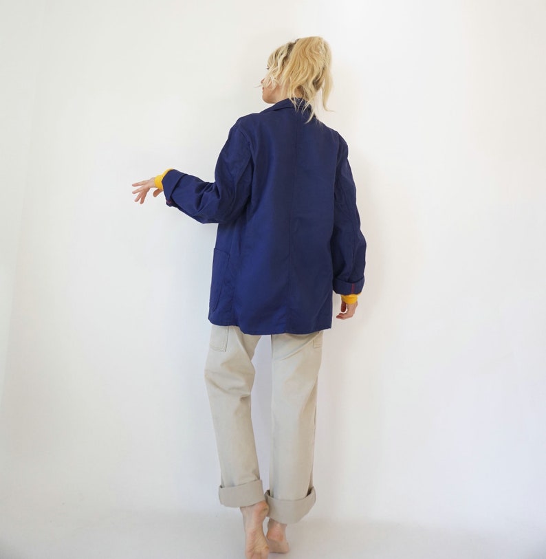 Pu&ograve; includere: Un blazer blu navy con due tasche anteriori e una manica gialla che sbuca. Il blazer &egrave; abbinato a pantaloni color cachi con risvolti. La persona &egrave; in piedi davanti a uno sfondo bianco.
