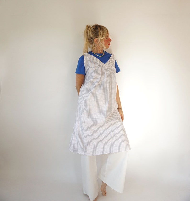 May include: A white and brown striped sleeveless dress with a V-neckline. The dress is worn over a blue t-shirt and white trousers.