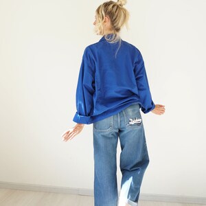 May include: A cobalt blue long-sleeve shirt and light wash denim jeans. The jeans have a white embroidered design on the back pocket. The jeans are cuffed at the ankles. The model has blonde hair in a bun.