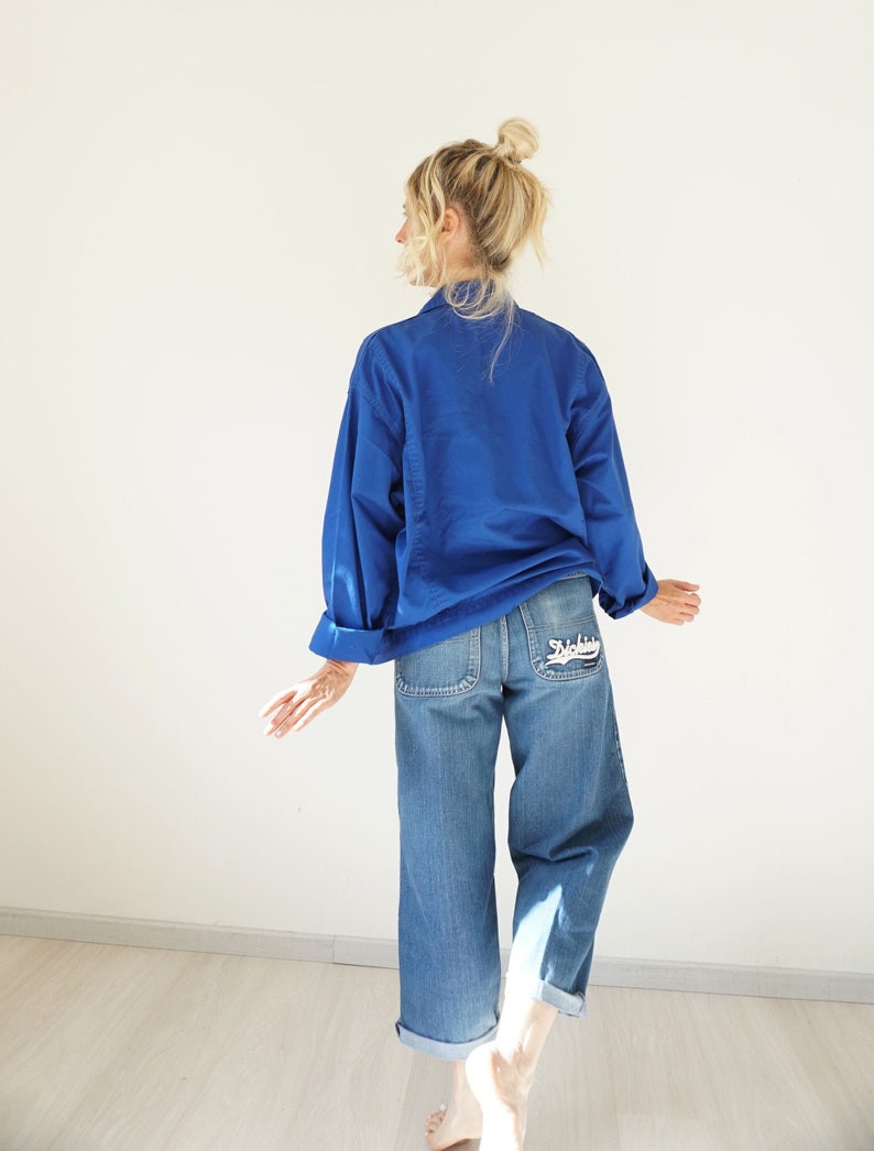May include: A cobalt blue long-sleeve shirt and light wash denim jeans. The jeans have a white embroidered design on the back pocket. The jeans are cuffed at the ankles. The model has blonde hair in a bun.