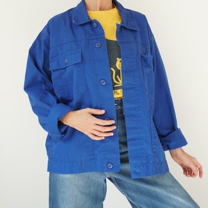 May include: A vibrant blue jacket with four front pockets and button closures. The jacket is worn over a yellow shirt and a graphic tee. The sleeves are cuffed, and the jacket is paired with blue jeans.