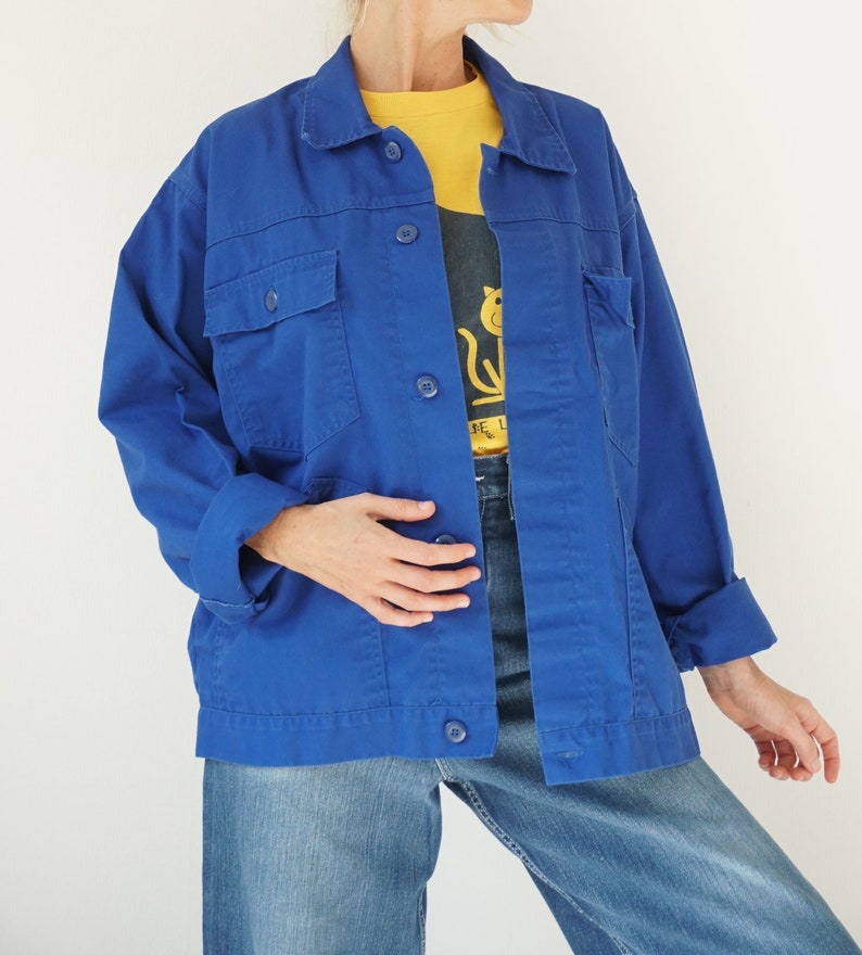 May include: A vibrant blue jacket with four front pockets and button closures. The jacket is worn over a yellow shirt and a graphic tee. The sleeves are cuffed, and the jacket is paired with blue jeans.