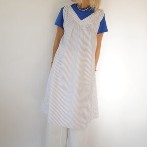 May include: A white sleeveless dress with thin blue stripes. The dress has a V-neck and a gathered bodice. The model is wearing the dress over a blue t-shirt and white trousers.