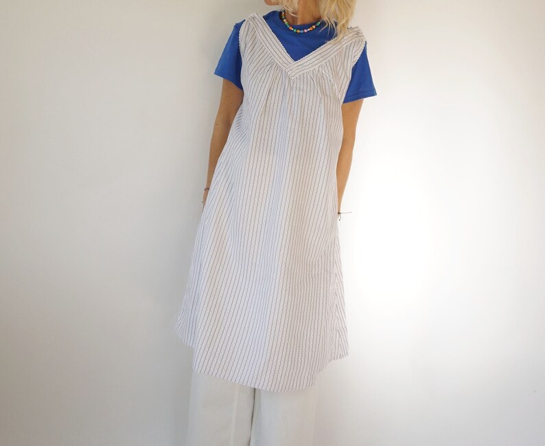 May include: A white sleeveless dress with thin blue stripes. The dress has a V-neck and a gathered bodice. The model is wearing the dress over a blue t-shirt and white trousers.