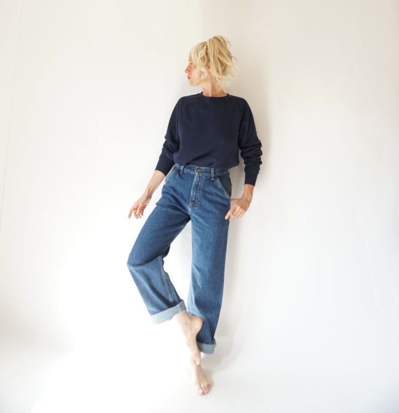 May include: A navy blue sweatshirt and blue denim jeans. The jeans have a classic fit with a folded hem. The model is standing on one foot against a white background. The sweatshirt has a crew neck.