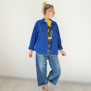 May include: A woman wearing a bright blue jacket over a yellow t-shirt and blue jeans. The jacket has two chest pockets and button closures. The jeans are wide-legged and cuffed at the ankles. The t-shirt has a graphic design.