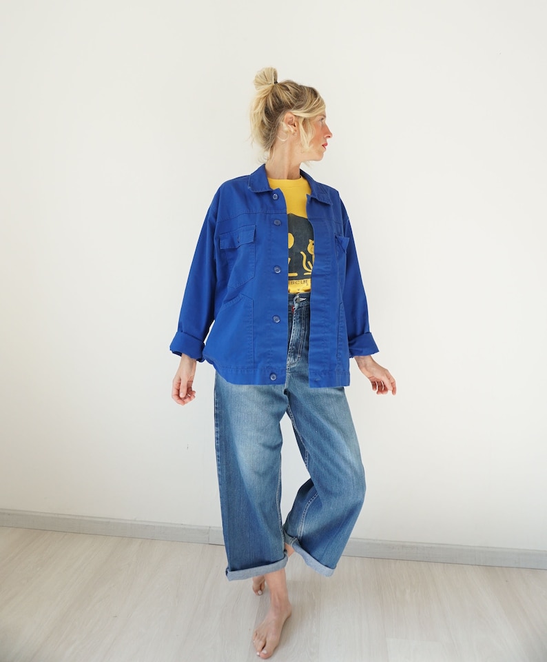 May include: A woman wearing a bright blue jacket over a yellow t-shirt and blue jeans. The jacket has two chest pockets and button closures. The jeans are wide-legged and cuffed at the ankles. The t-shirt has a graphic design.
