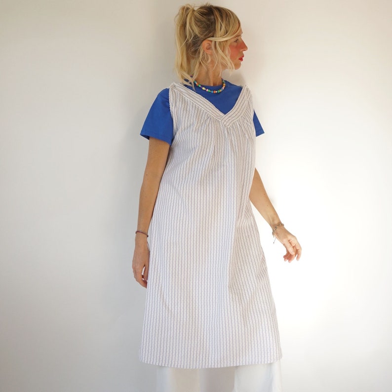 May include: A white and blue striped sleeveless dress with a V-neckline. The dress is worn over a blue t-shirt.