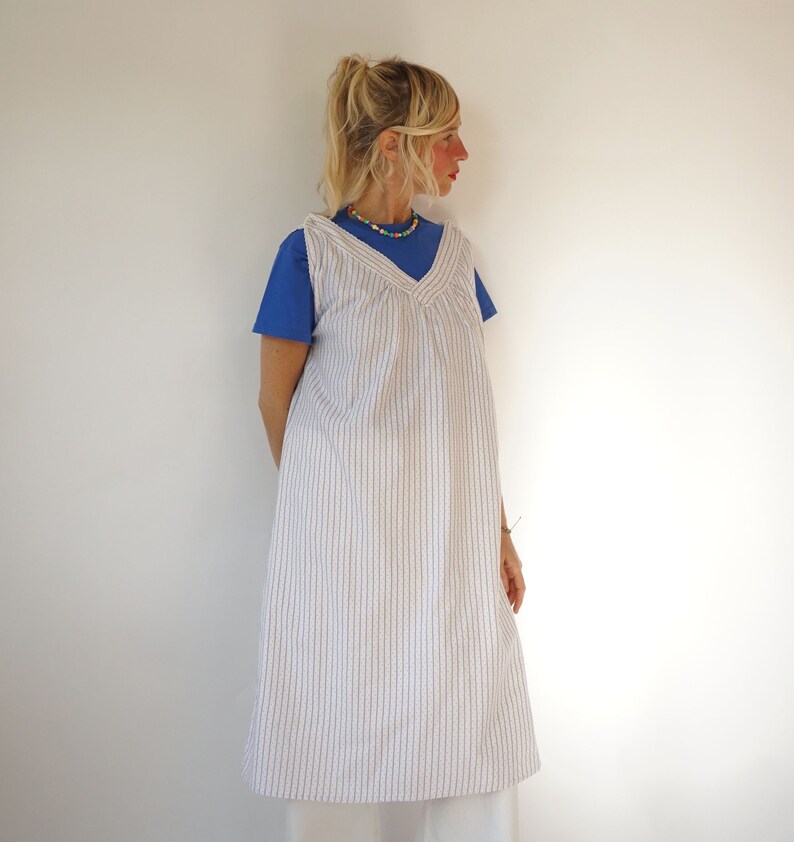 May include: A white and blue striped sleeveless dress with a V-neckline. The dress is worn over a blue t-shirt. The model is wearing a colourful beaded necklace.