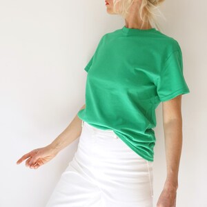 May include: A solid green t-shirt with a crew neck. The shirt is paired with white pants. The model is wearing the shirt and pants. The background is a plain white wall.