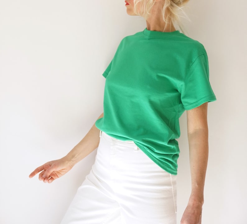 May include: A solid green t-shirt with a crew neck. The shirt is paired with white pants. The model is wearing the shirt and pants. The background is a plain white wall.