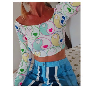 May include: A white long-sleeved crop top with a colorful yin-yang pattern featuring hearts. The top has an off-the-shoulder neckline and is made of a stretchy fabric.