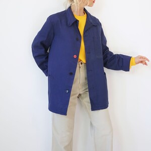 Pu&ograve; includere: Giacca da lavoro blu navy con chiusura a bottoni e una tasca sul petto. La giacca ha un colletto classico e maniche lunghe. I bottoni sono neri e rossi. La modella indossa una camicia gialla e pantaloni beige.
