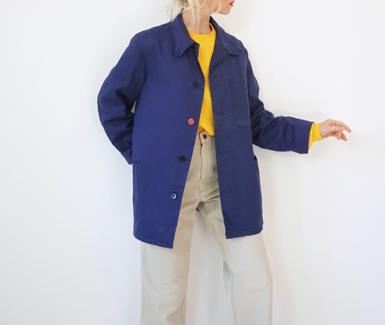 Pu&ograve; includere: Giacca da lavoro blu navy con chiusura a bottoni e una tasca sul petto. La giacca ha un colletto classico e maniche lunghe. I bottoni sono neri e rossi. La modella indossa una camicia gialla e pantaloni beige.